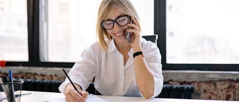 Smiling woman talking on the phone while writing notes at a desk in a modern office.